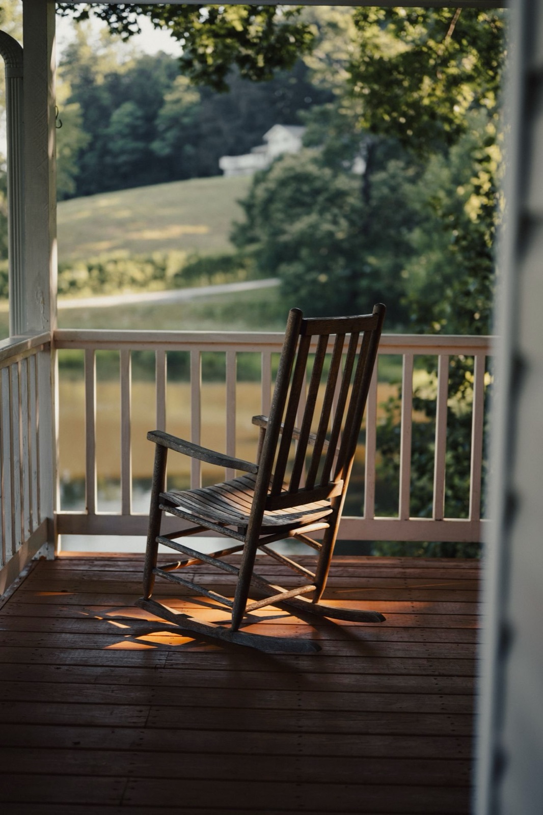 An old wooden rocking chair on the porch of a country house, warm light across the floorboards and green hills beyond.