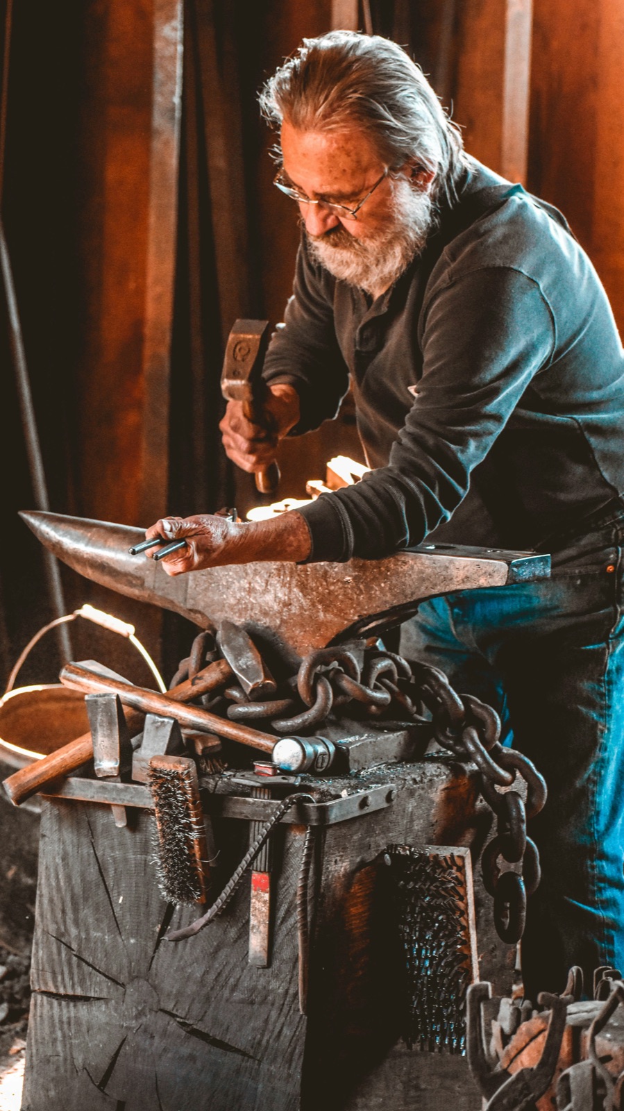 A blacksmith at the forge holding a hammer, working glowing iron in the dim light of the shop.