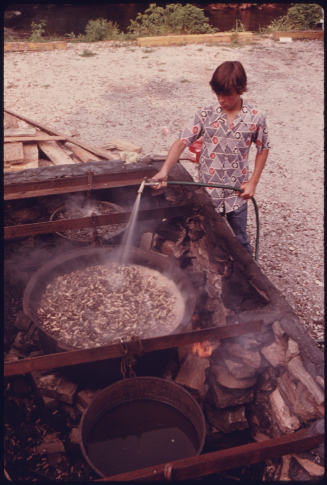 A child sprays water from a hose into a large open pan of peanuts boiling over a wood fire on a gravel lot.