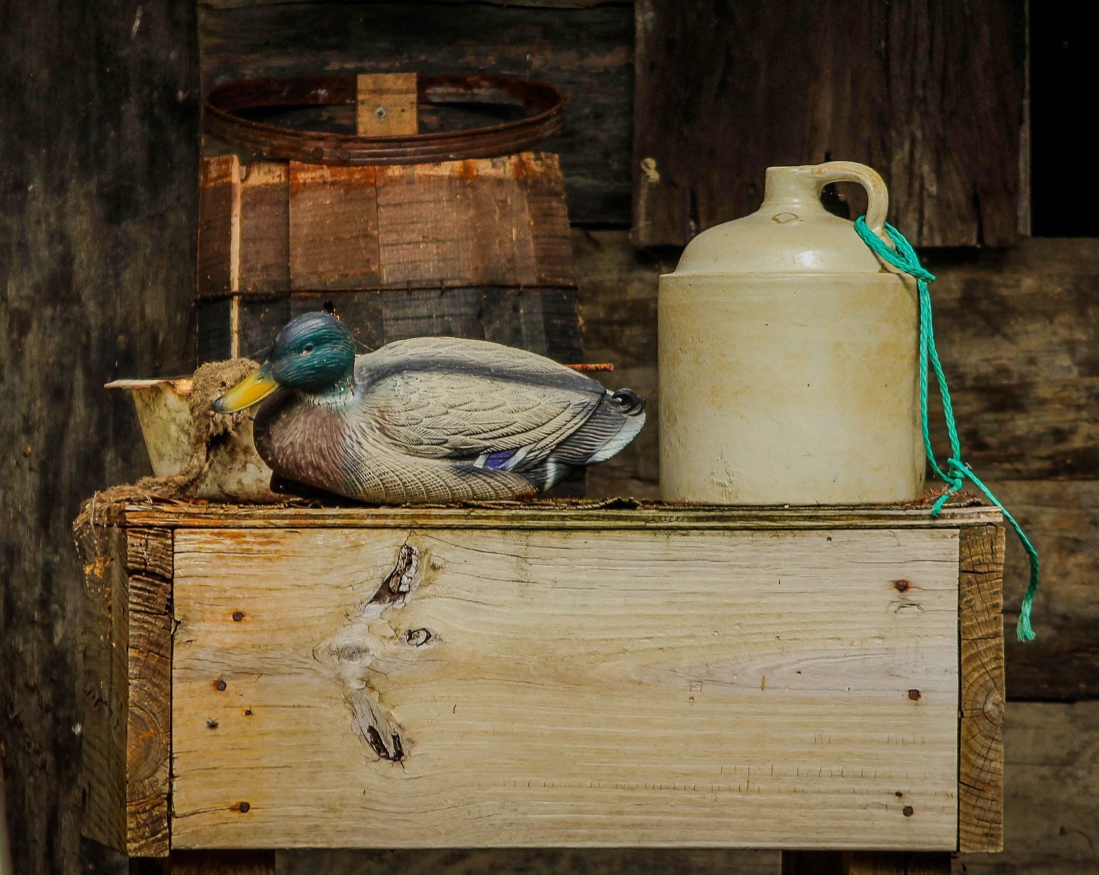 A painted wooden mallard decoy resting on burlap atop a rough wooden crate, beside an old stoneware jug against weathered barn boards.