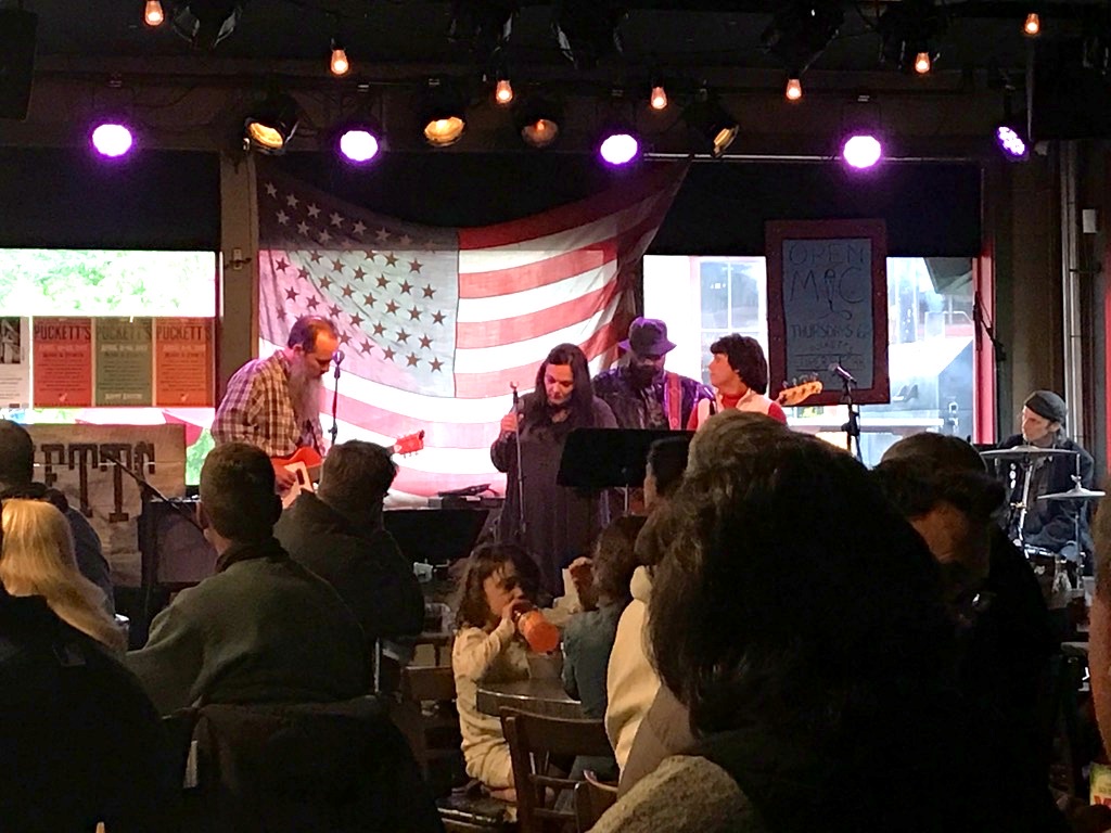 An open-mic night at the Leiper's Fork grocery, a band on stage beneath a large American flag.