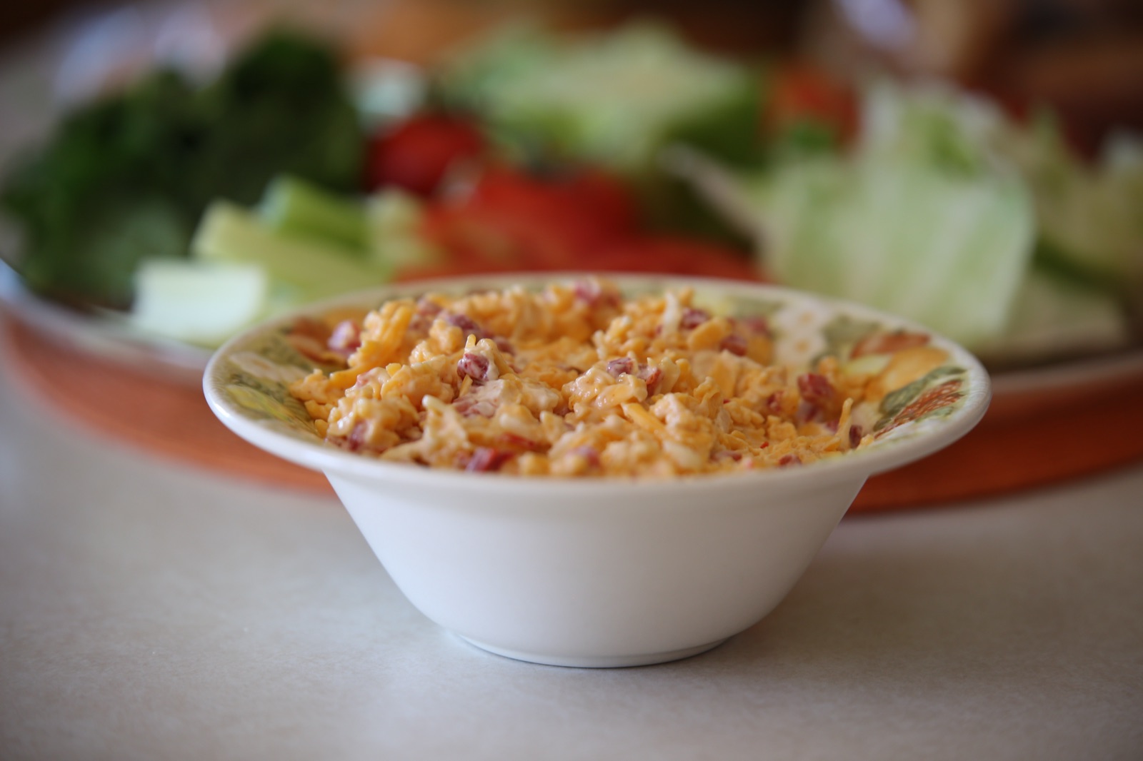 A white bowl of homemade pimento cheese with fresh vegetables behind it.