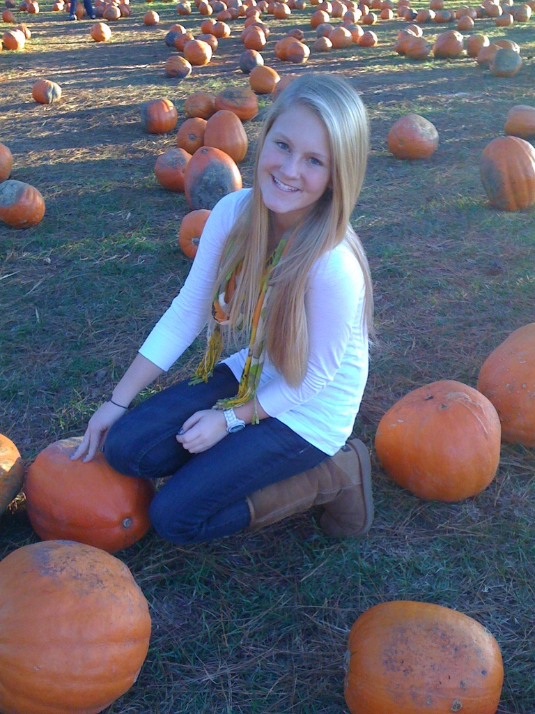 A young woman in a white top and yellow scarf kneels smiling among big orange pumpkins in a sunny pumpkin patch.