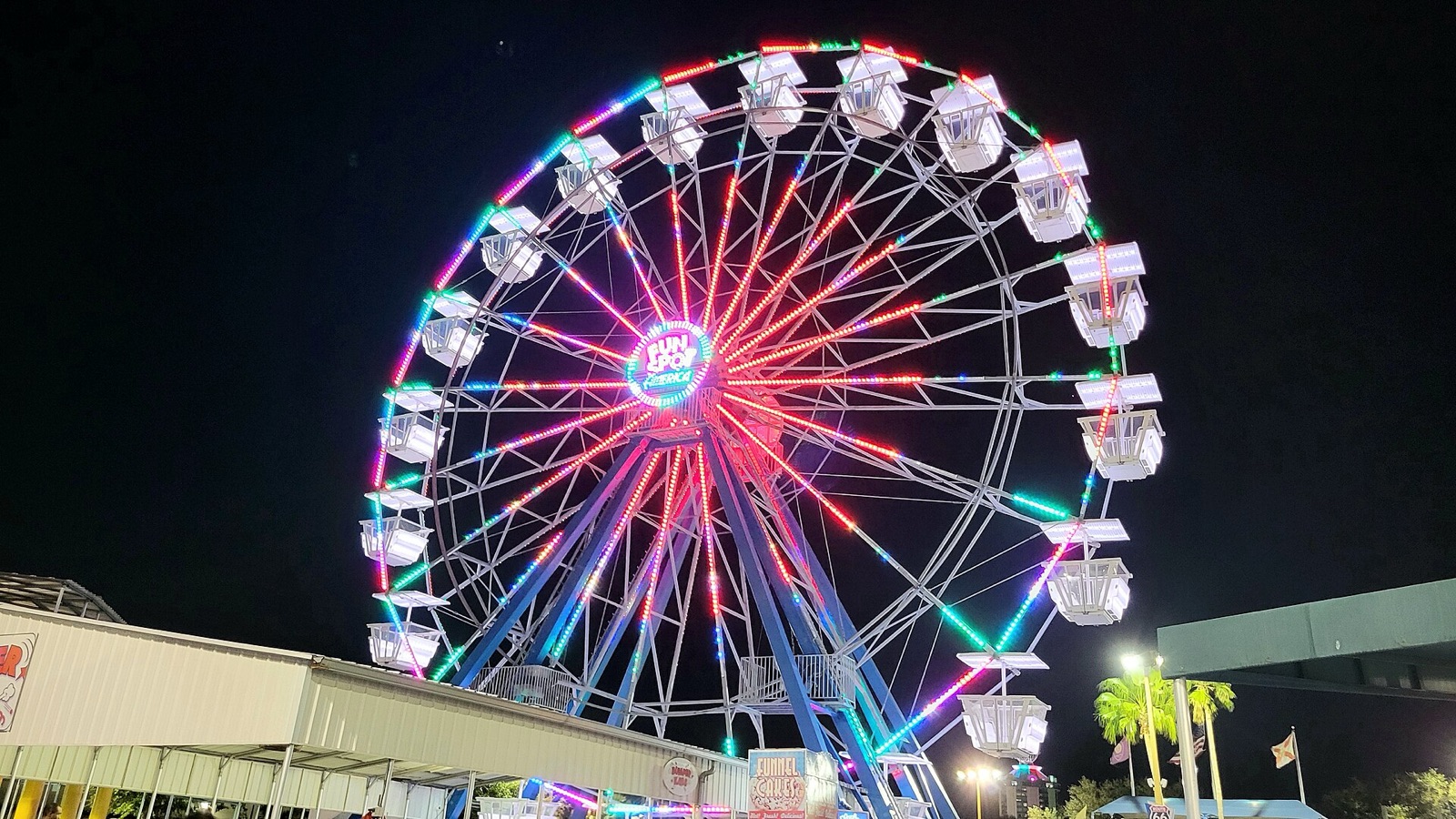 A Southern county fair midway glowing at dusk, a lit Ferris wheel turning above the crowd and game booths.
