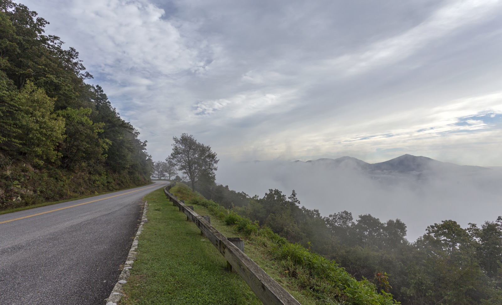 The Blue Ridge Parkway curving along a forested mountainside above a valley of low clouds.
