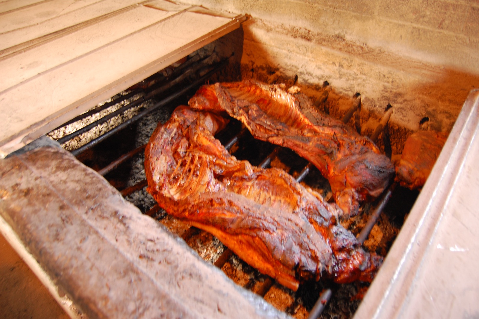 A whole hog browning on a metal grate inside an open brick barbecue pit, cooking low over coals and smoke.